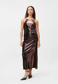 Sequined evening dress in dark bronze with a front slit and spaghetti straps; smooth texture and fitted silhouette. Black heels accent the look.