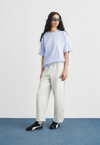 Ανοιχτό μωβ βαμβακερό μπλουζάκι με λογότυπο, γκρι φόρμα με λεπτομέρειες της μάρκας και μαύρα αθλητικά παπούτσια. Τοποθετημένα σε απλό φόντο.