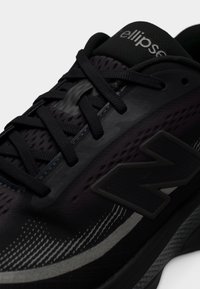 Černá atletická obuv s horní částí z prodyšné síťoviny, texturovanými akcenty a reflexními detaily. Má výrazné šedé logo a černé tkaničky.