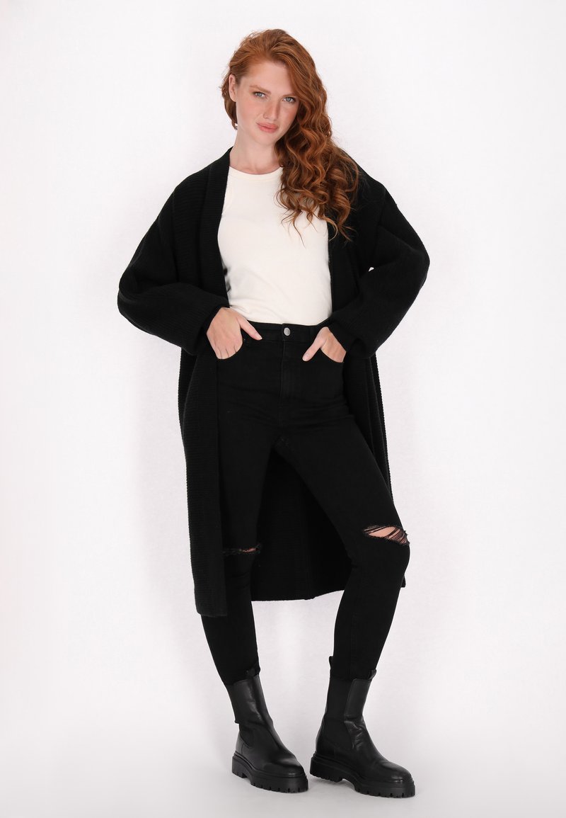 Zwarte lange cardigan over een witte top, gecombineerd met hooghals zijs zwart jeans met gescheurde knieën en zwarte enkellaarsjes.