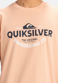 Camiseta de algodón melocotón con un logotipo gráfico en blanco y negro de Quiksilver que incluye el texto "THE ORIGINAL BOARDRIDING CO" y un diseño de ola.