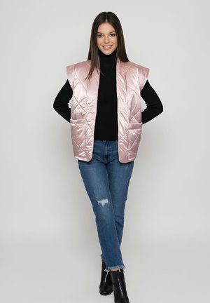 Joven mujer con un chaleco acolchado sin mangas de color rosa brillante, suéter de cuello alto negro, jeans azules rasgados y botines negros, de pie con las manos detrás de la espalda.