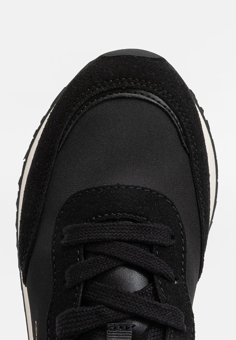 Svart sneaker med en texturerad matt ovandel, rund tå, platta snören och en kontrasterande vit mellansula med mörk gummisula.