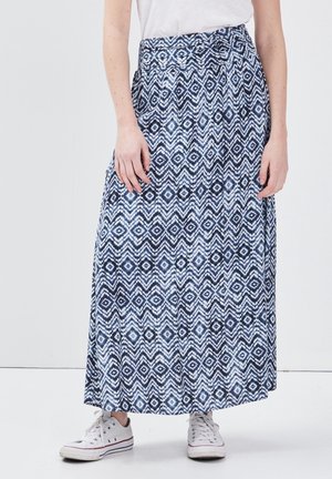 Persona che indossa una gonna lunga con motivo geometrico blu e bianco, sneakers bianche e una maglietta bianca, in piedi su uno sfondo uniforme.