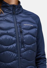 Marineblauwe geïsoleerde jas met een gewatteerde textuur, hoge kraag en ritszak aan de voorkant. Bevat het Peak Performance-logo op de borst.