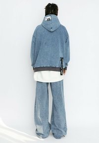 Sudadera de mezclilla azul claro de gran tamaño con detalles en gris y una etiqueta en el lateral, combinada con pantalones de mezclilla azul claro de pierna ancha.