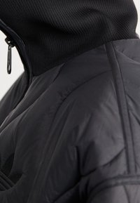 Chaqueta acolchada negra con alto cuello y cuello acanalado, que presenta una textura suave y detalles de costura. Cierre de cremallera y logo visible.