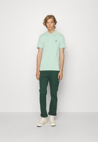 Muntgroene polo met een kraag en korte mouwen, met een klein geel logo op de borst, gecombineerd met donkergroene broeken en witte schoenen.