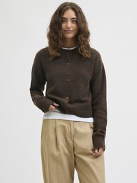 Jovem mulher com cabelo castanho ondulado, vestindo um cardigã castanho escuro com botões sobre uma camisa branca e calças bege, de pé contra um fundo plain.