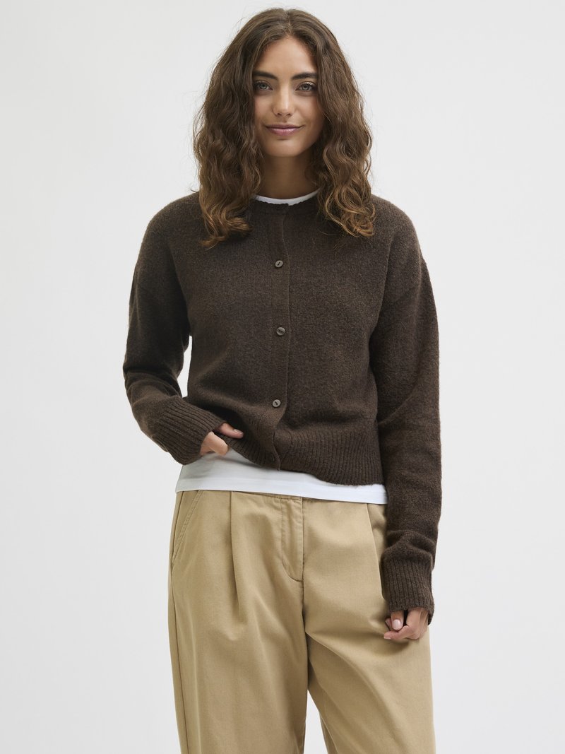 Jovem mulher com cabelo castanho ondulado, vestindo um cardigã castanho escuro com botões sobre uma camisa branca e calças bege, de pé contra um fundo plain.