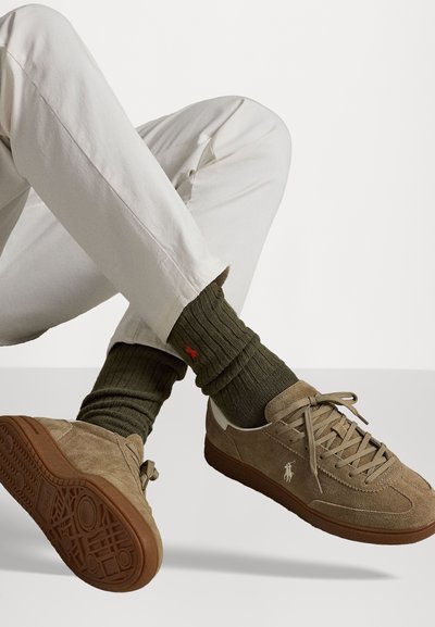 Zapatillas de ante marrón con un upper texturizado, cordones y suela de goma, combinadas con calcetines de punto verdes oscuros que presentan un logo de acento.