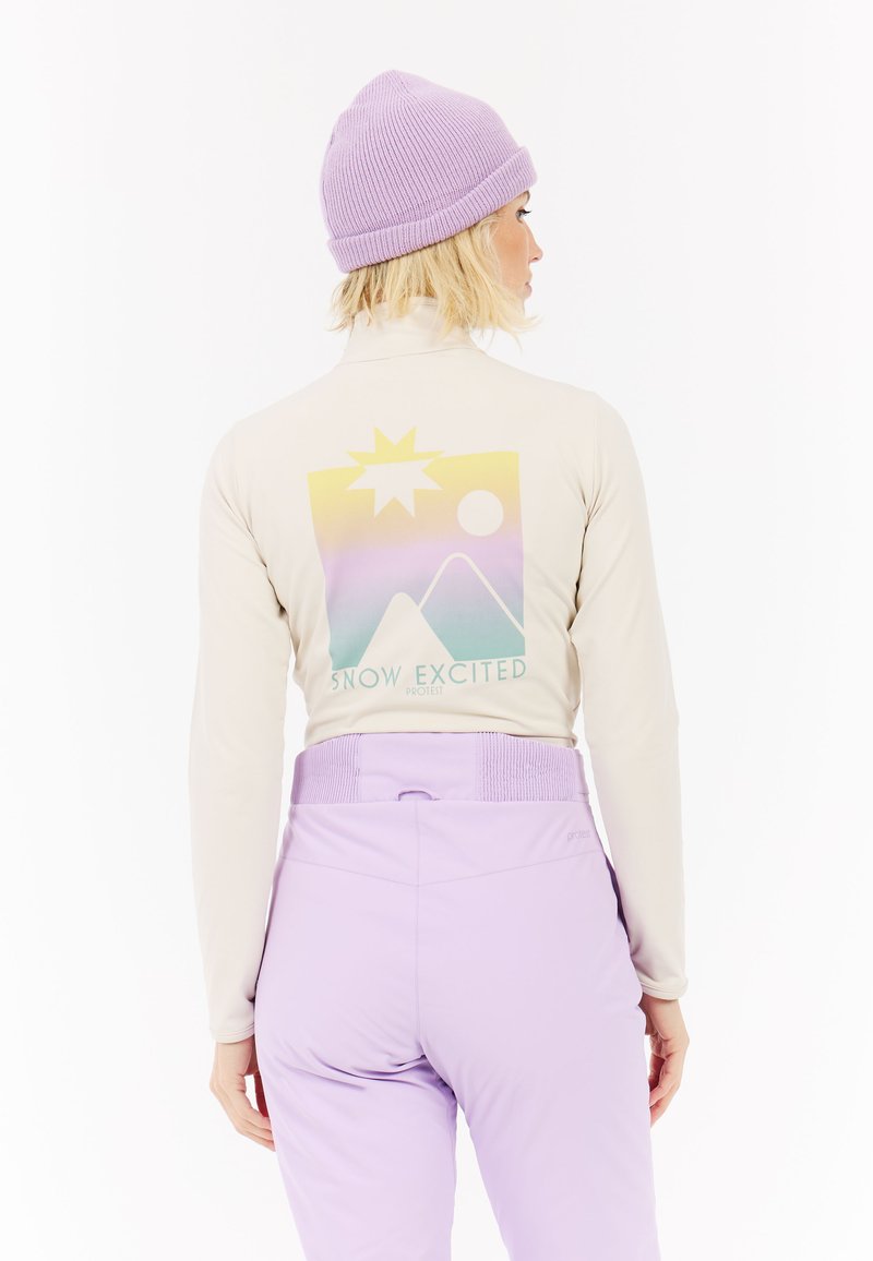 Lange mouwen crèmekleurig bovenstuk met een pastel bergen- en zongrafiek op de achterkant, gecombineerd met lichtpaarse skibroek en een lavendelkleurige gebreide beanie.