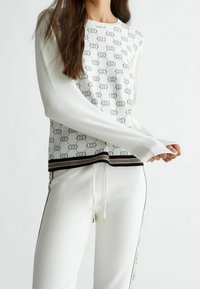 Pullover bianco a maglia con motivo del logo ripetuto, polsini a costine e orlo a strisce nere e oro. Abbinato a pantaloni bianchi con cordino.