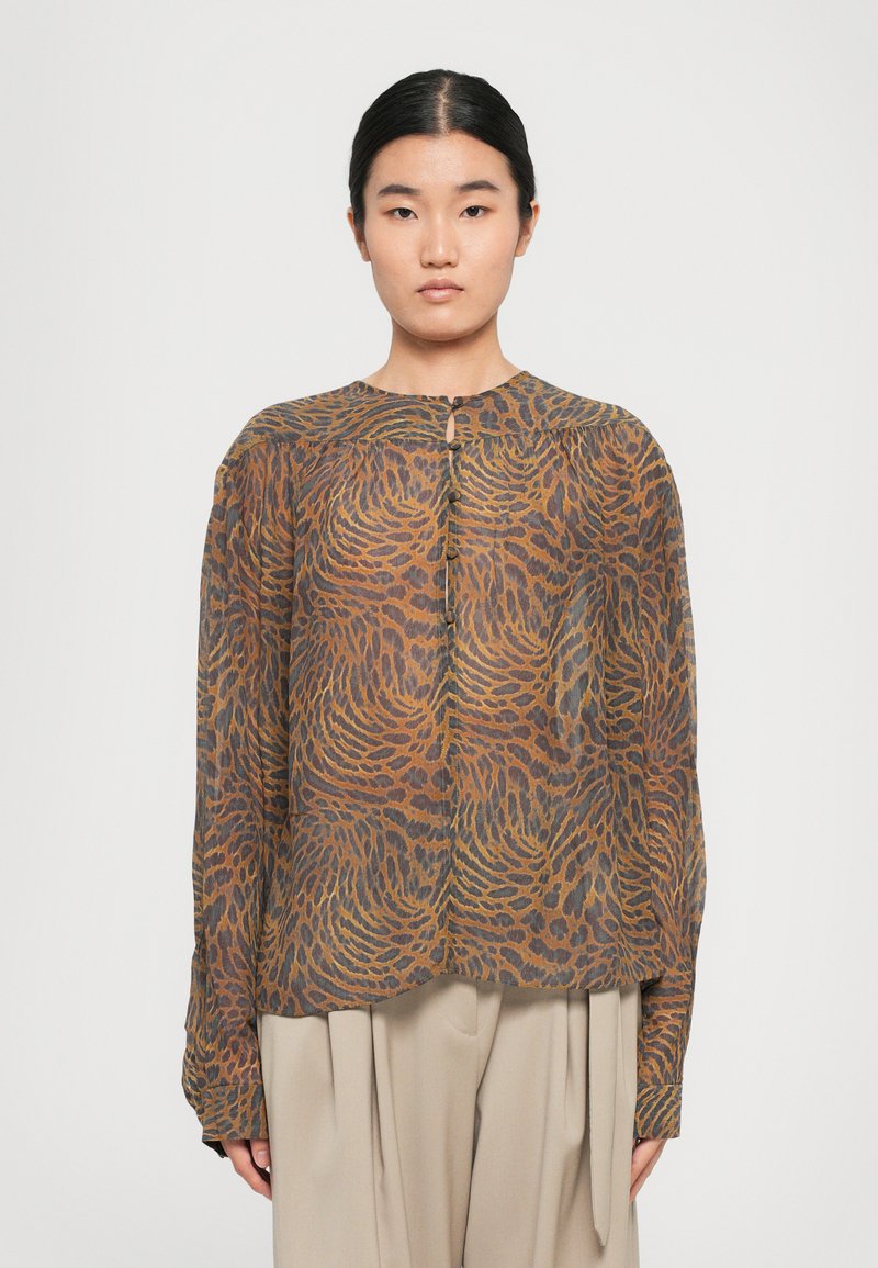 Žena oblečená v hnedom patterned blouse s dlhým rukávom a gombíkmi, s béžovými širokými nohavicami, stojí pred jednoduchým bielym pozadím.