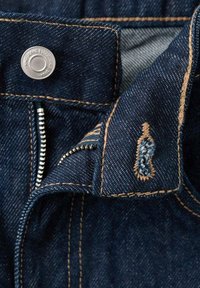Temno modra jeans tkanina z oranžnim šivom, z nastavljanjem na kovinsko gumb in zadrgo. Vidna je dekorativna modra vezenina.