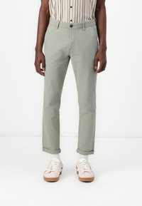 Pantaloni in cotone verde chiaro con risvolti, dal taglio standard e tasche laterali, indossati con sneakers beige e calzini bianchi.