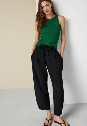 Femeie care stă în picioare, purtând un top verde fără mâneci cu detalii florale, pantaloni largi negri cu talie cu panglică și sandale negre tip flip-flop.