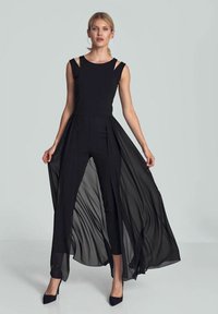 Zwarte jumpsuit met transparante, vloeiende overlay. Bevat schouderuitsnijdingen, een getailleerde buste en een uitlopende onderkant. Gladde textuur en elegante lijnen.