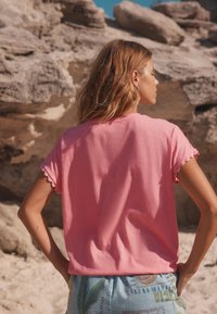 Rózsaszín, rövid ujjú póló fodros ujjakkal, könnyű anyagból, mintás világoskék rövidnadrággal párosítva. Természetes sziklás háttér.