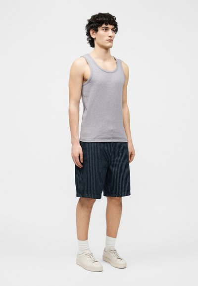 Νεαρός άνδρας που στέκεται, φοράει γκρι αμάνικο μπλουζάκι, σορτς με μπλε ναυτικό χρώμα και λεπτές ρίγες, λευκές κάλτσες και μπεζ αθλητικά παπούτσια σε απλό φόντο.