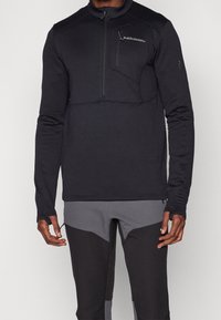 Zwarte quarter-zip pullover gemaakt van gestructureerde stof, met een borstzak en duimgaten in de mouwen. Draagt met zwarte en grijze broek.