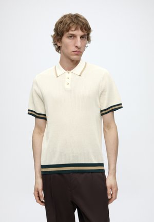 Jeune homme aux cheveux bouclés portant un polo en maille crème avec des bordures rayées vertes et beiges, debout devant un fond uni clair.