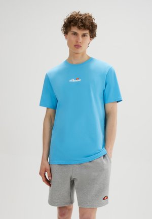 Joven con pelo rizado que lleva una camiseta azul de ellesse y pantalones cortos grises de ellesse, de pie frente a un fondo liso y claro.