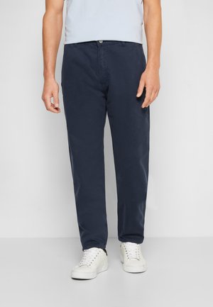 Mann trägt marineblaue Hose mit geradem Bein und weißen Sneakers, steht vor einem einfachen hellgrauen Hintergrund.