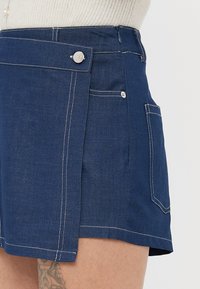 Marineblå denimshorts med hvite sømmer, med en knapp på siden, frontlomme og et unikt brettedesignelement.