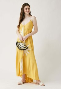 Gelbes Maxikleid mit Spaghetti-Trägern, vorderen Knöpfen und Blumenstickerei. Kombiniert mit einer schwarz-weißen Perlenhandtasche und flachen Sandalen.