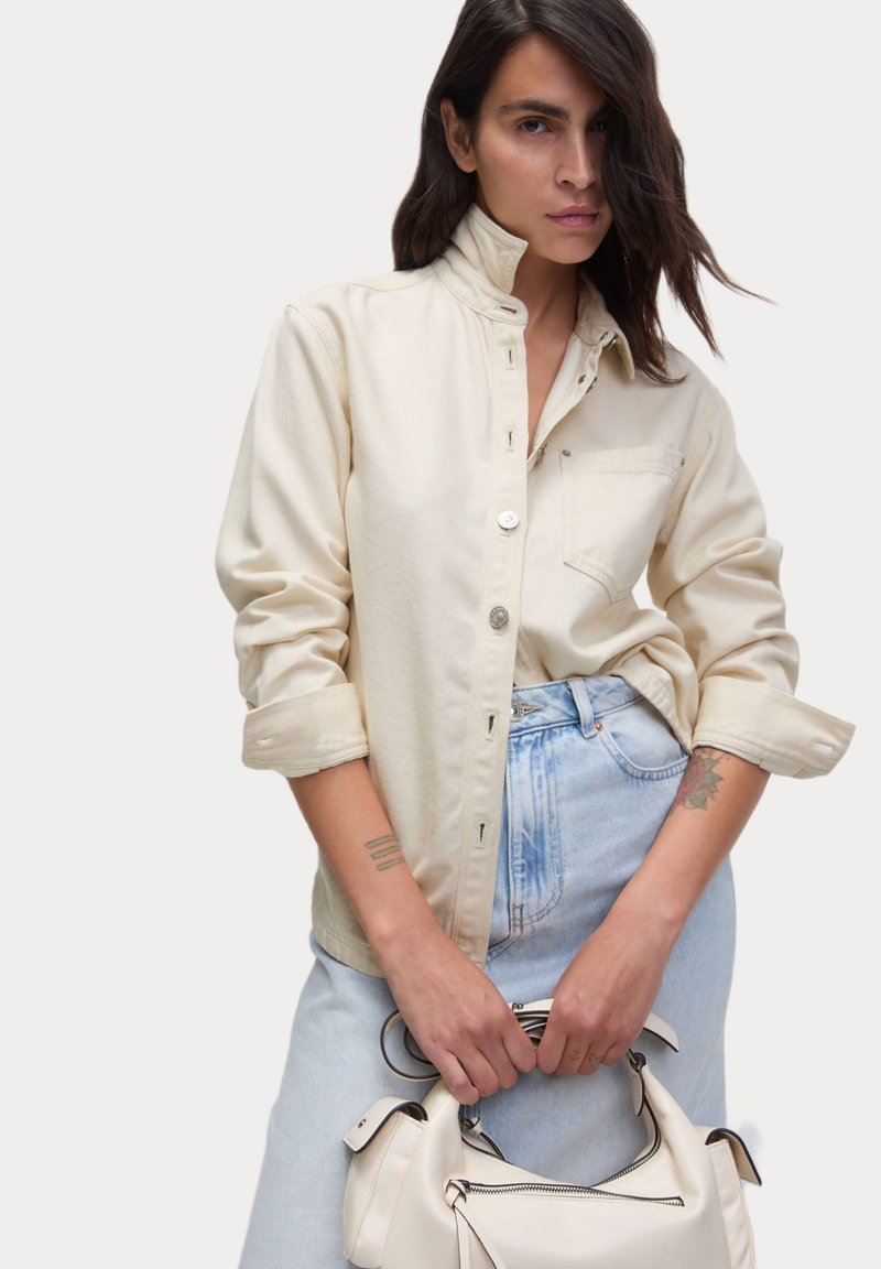 Lichtbeige overhemd metopstaande kraag, lange mouwen en een voorzak, gecombineerd met lichtblauwe denim en een witte handtas.