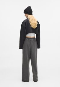 Blonde oseba, oblečena v črno kapo, skrajšan črn pulover, bel top in ohlapne sive hlače, stoji obrnjena stran od kamere na enobarvni beli podlagi.