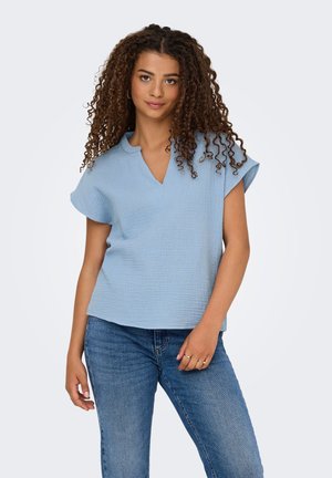 Jonge vrouw met krullend haar, die een lichtblauw gestructureerd kort mouwen blouse en blauwe spijkerbroek draagt, staand tegen een effen achtergrond.