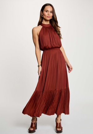 Donna in piedi con un abito lungo plissettato senza maniche color ruggine, collo alto e sandali marroni con tacco su sfondo neutro.