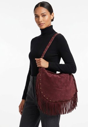 Femme en col roulé noir tenant un grand sac à épaule en daim bordeaux avec franges et clous en laiton.