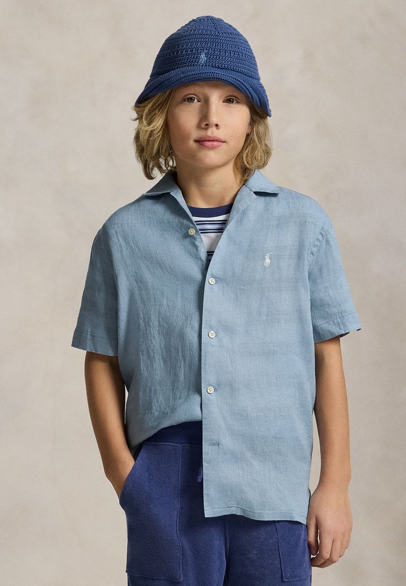 Enfant aux cheveux blonds mi-longs portant un chapeau seau bleu en maille, une chemise bleu clair à manches courtes boutonnée, avec un t-shirt rayé bleu marine en dessous.
