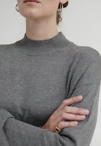 Grauer Rollkragenpullover aus weichem, geripptem Strickstoff; verfügt über lange Ärmel und eine lockere Passform, mit einer Nahaufnahme des Kragens und gefalteten Händen.