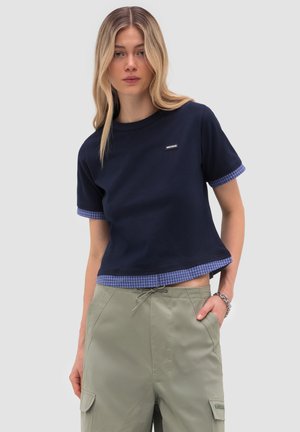 Mujer joven con una blusa azul marino de manga corta con ribete de cuadros azules y pantalones cargo verde claro, de pie con una mano en el bolsillo.