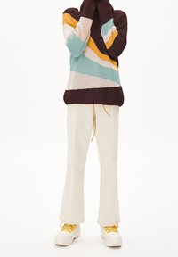 Pull en tricot multicolore avec des rayures marron, sarcelle, jaune et crème ; associé à un pantalon évasé crème et des baskets blanches avec des lacets jaunes.