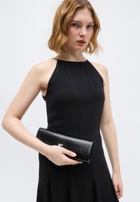 Νεαρή γυναίκα με αμάνικο μαύρο φόρεμα κρατώντας μια μαύρη δερμάτινη τσάντα χειρός μπροστά σε ένα απλό ανοιχτό φόντο.