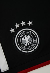 Čierna džersejová látka s okrúhlou vyšitú nášivkou s dvojitým kruhom, textom "DEUTSCHER FUSSBALL-BUND" a štyrmi bielymi hviezdami nad ňou.