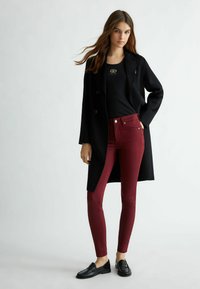 Cappotto lungo nero, maglione aderente nero, jeans skinny bordeaux e mocassini neri. Design semplice con accento del logo metallico sul maglione.
