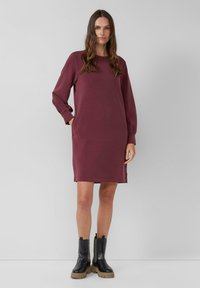 Bordeauxfarbenes Sweatshirtkleid mit langen Ärmeln, Rundhalsausschnitt und seitlichen Taschen. Kombiniert mit schwarzen Ankle Boots und einer klobigen, braunen Sohle. Glatte Textur.