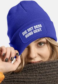 Blue knit beanie with a textured pattern and a rectangular patch featuring white text. The design is simple and functional.