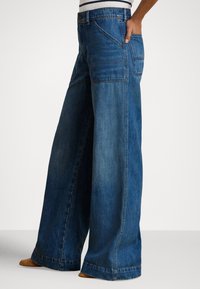 Vide blå jeans med høy midje, vide ben, lommer med lapper, vasket utseende og frynsete kanter. Passer til uformell bruk.