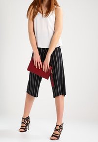 Mujer vistiendo una blusa sin mangas blanca y culottes a rayas negras, sosteniendo un bolso de mano rojo, de pie en zapatos de tacón alto negros con tiras.