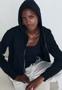 Zwarte zip-up hoodie met een capuchon, gladde stof, gouden rits en geribbelde manchetten. Gekocht over een zwarte tanktop en gecombineerd met lichte, gemusterde broeken.