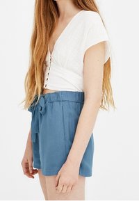 Top bianco a maniche corte con bottoni frontali, abbinato a pantaloni corti blu con coulisse caratterizzati da una texture liscia e una vestibilità comoda.