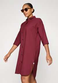 Vestido burdeos de botones con mangas tres cuartos, aberturas laterales y botones negros; tejido suave con un corte relajado.