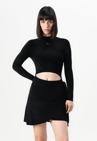 Čierny top s dlhým rukávom a vykrojeným pásom, v páre s zhodnou zavinovacou sukňou. Tkanina má jemnú štruktúru a hladký povrch, minimalizované detaily.